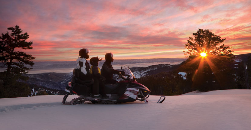 Three people on a snowmobile looking out to the setting sun