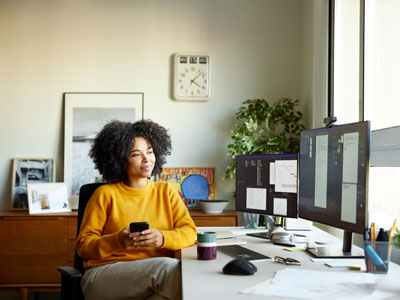 woman in a gold top holding a cell phone, looking at computer screens