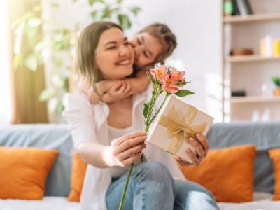 Mother holding a gift box and flowers and a little girl behind her with her arms wrapped around the mothers neck