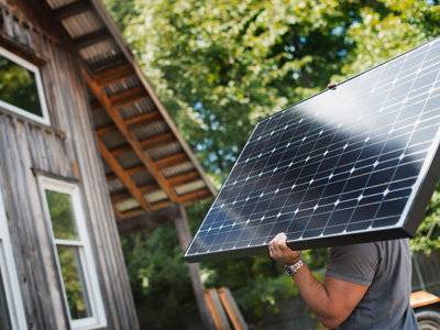 A person carrying a solar panel toward a house