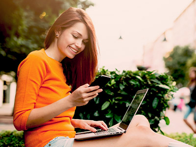 Woman in orange looking at phone with laptop on lap while sitting on grass