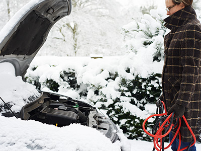 a person holding cables looking at a car covered in snow with an opened hood.