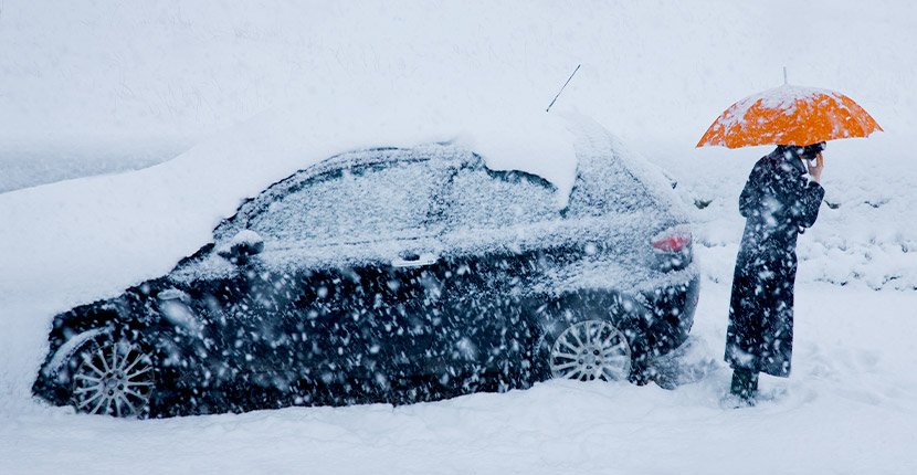 Person standing in the snow under an umbrella next to the back of a black car